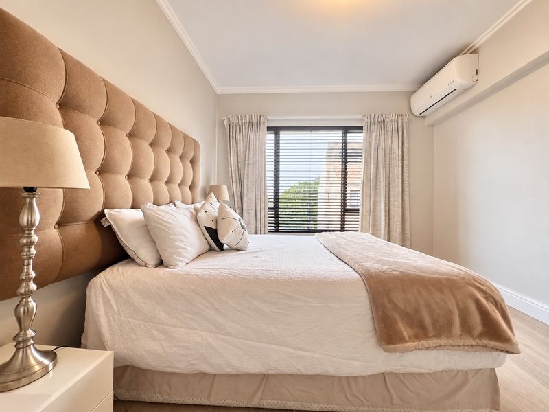 A well-maintained bedroom featuring neutral tones, laminate flooring, a wall-mounted air conditioning unit, and window treatments including venetian blinds and curtains.