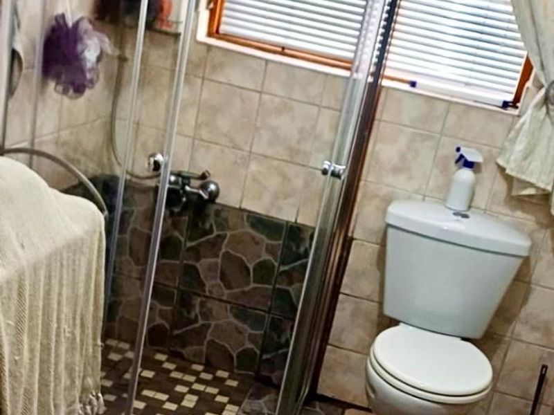 A functional bathroom featuring a corner shower enclosure with stone-look tiling, a standard toilet, and a window with fitted blinds.