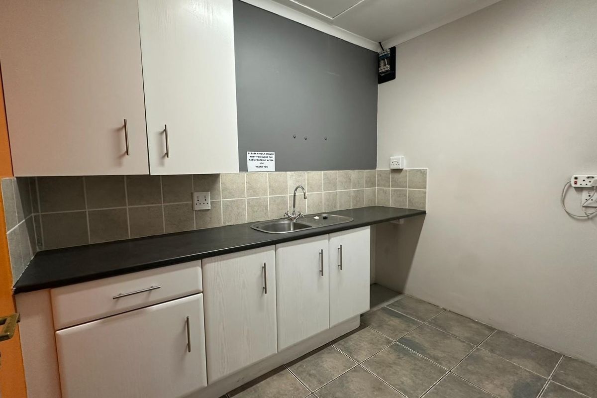 A functional office kitchenette featuring white cabinetry, a dark countertop, a stainless steel sink, and tiled splashbacks.