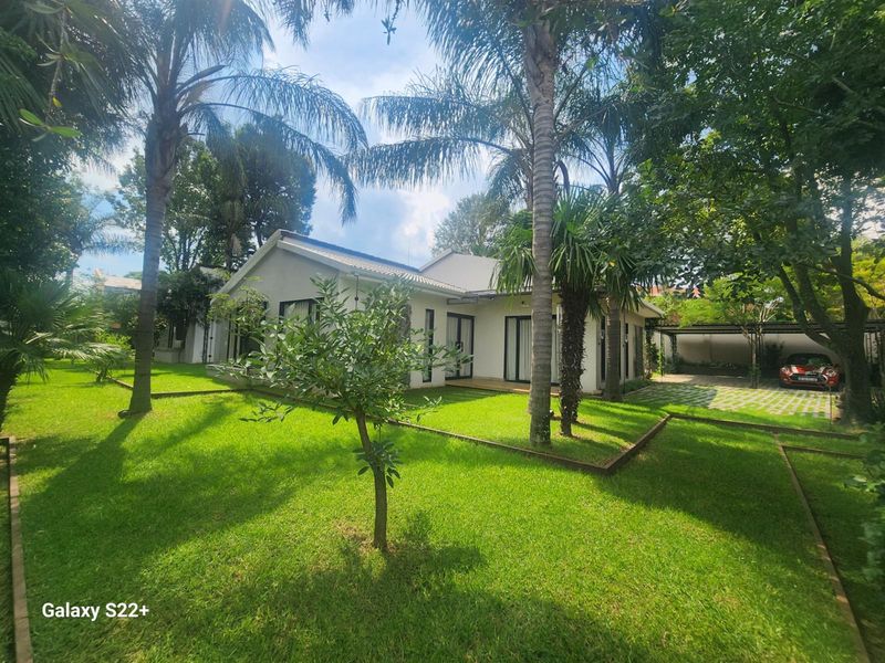 A well-maintained rear exterior view of the house featuring a lush green garden, mature palm trees, and a paved driveway with a carport.