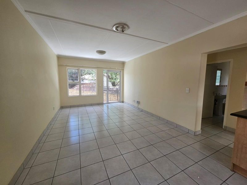An empty, open-plan living area featuring neutral tiled flooring, large windows, and a sliding glass door providing access to the exterior.