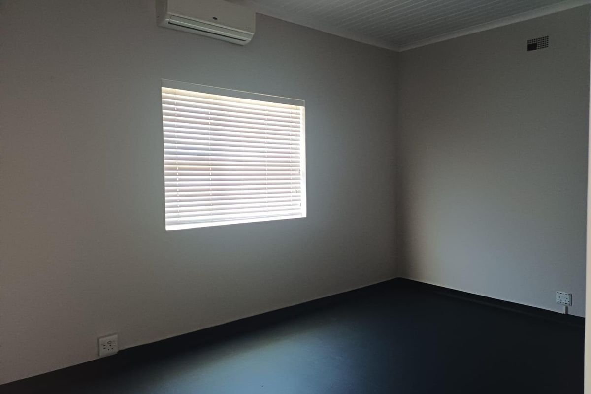 A clean, empty bedroom featuring a wall-mounted air conditioning unit, venetian blinds, and dark flooring.
