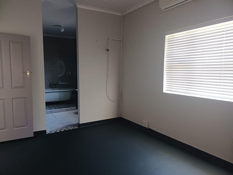 An empty bedroom featuring dark flooring, a window with fitted blinds, and an adjoining bathroom with a freestanding bathtub.