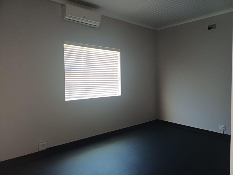 A clean, empty bedroom featuring a wall-mounted air conditioning unit, venetian blinds, and dark flooring.