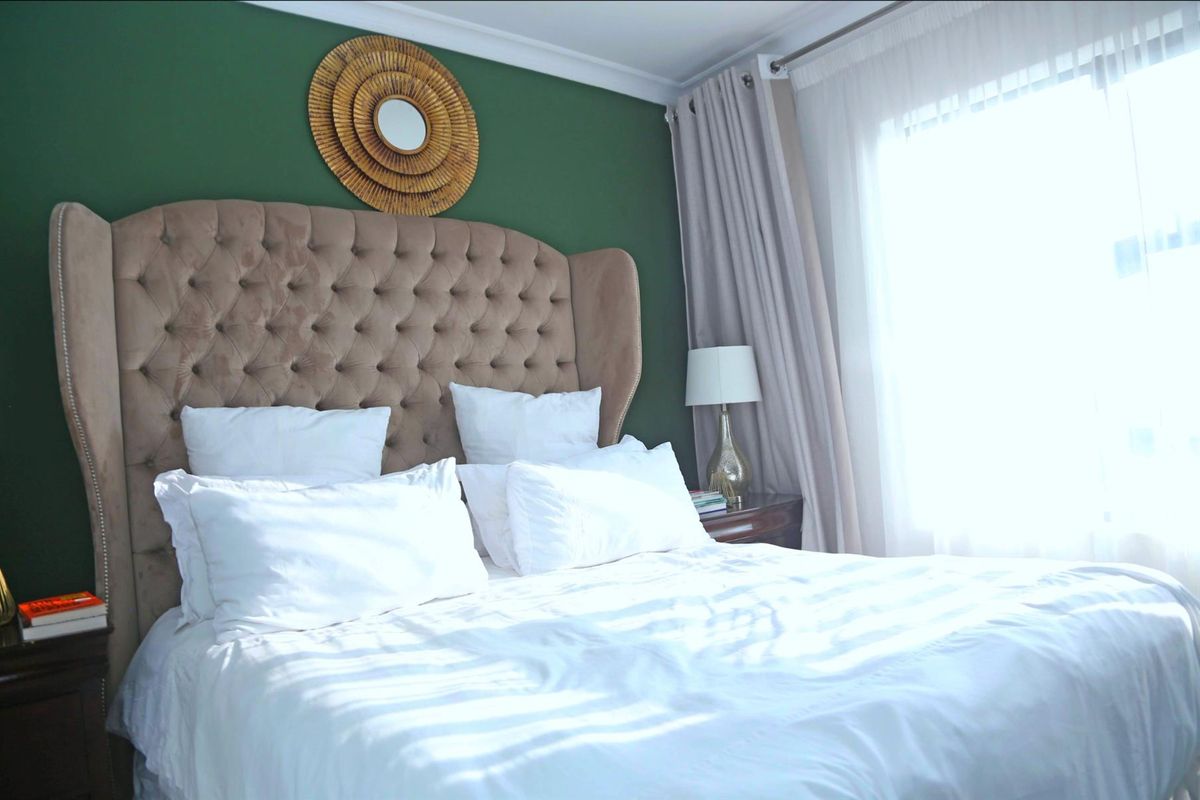 A well-maintained bedroom featuring a bold green accent wall, large window with natural light, and neutral carpeting.