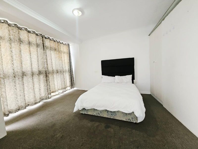 A spacious bedroom featuring neutral walls, built-in cupboards, and large windows with floor-to-ceiling curtains, providing ample natural light.