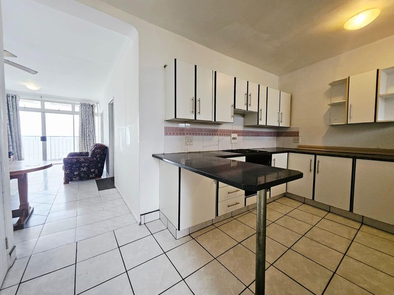 Open-plan kitchen featuring white cabinetry, dark countertops, a breakfast bar, and tiled flooring, seamlessly flowing into the adjacent living area with balcony access.
