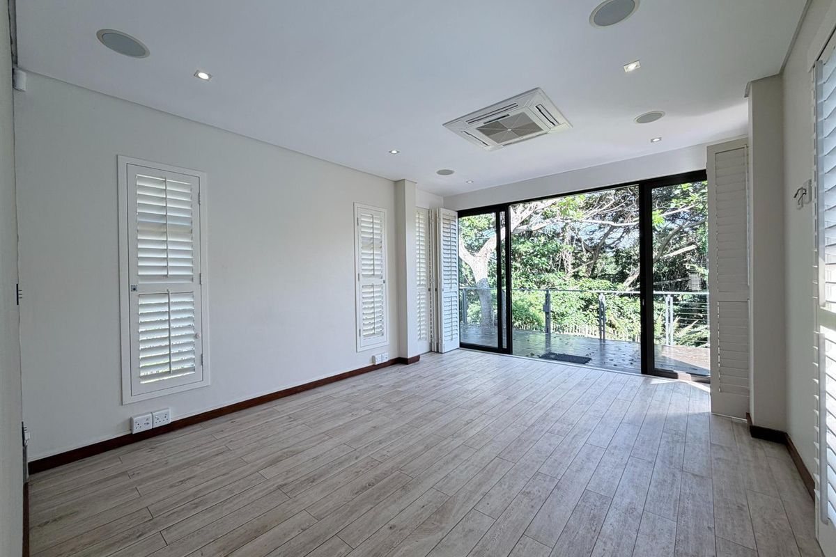 Bright living area featuring wood-look tile flooring, recessed ceiling speakers, integrated air conditioning, and plantation shutters, opening onto a private balcony with lush views.