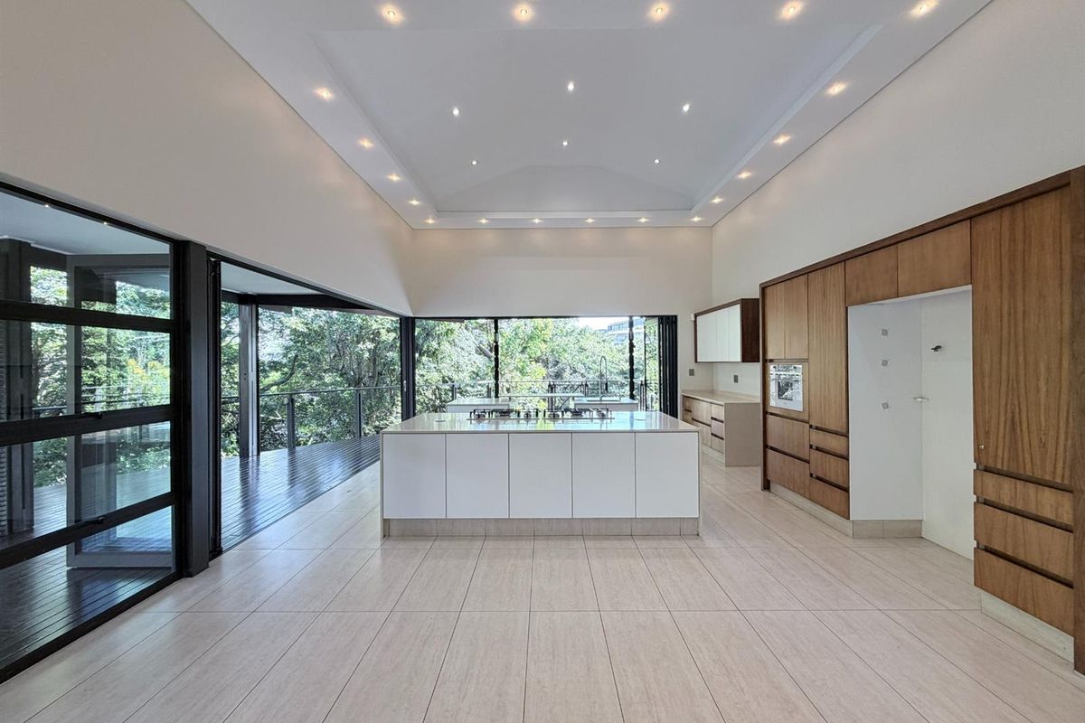 Spacious modern kitchen featuring a large central island with integrated cooktop, sleek wooden cabinetry, recessed ceiling lighting, and expansive glass doors opening to a deck.