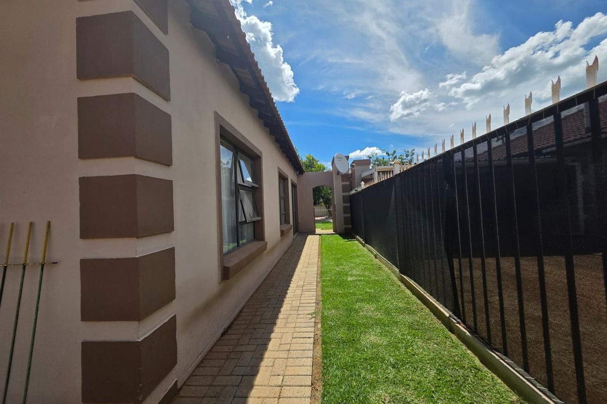 A side exterior view of the property featuring a paved walkway, a well-maintained lawn, and perimeter security fencing.