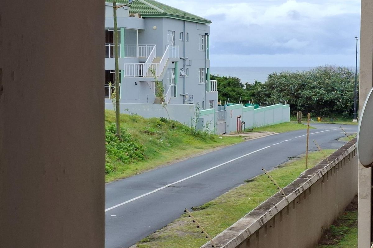 A street-level view showing the apartment complex exterior, surrounding road, and distant ocean view from a residential perspective.