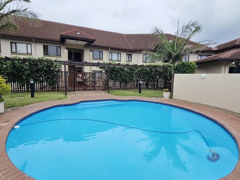 A communal swimming pool area featuring a paved surround, landscaped garden, and a wooden pergola, set against the apartment complex building.