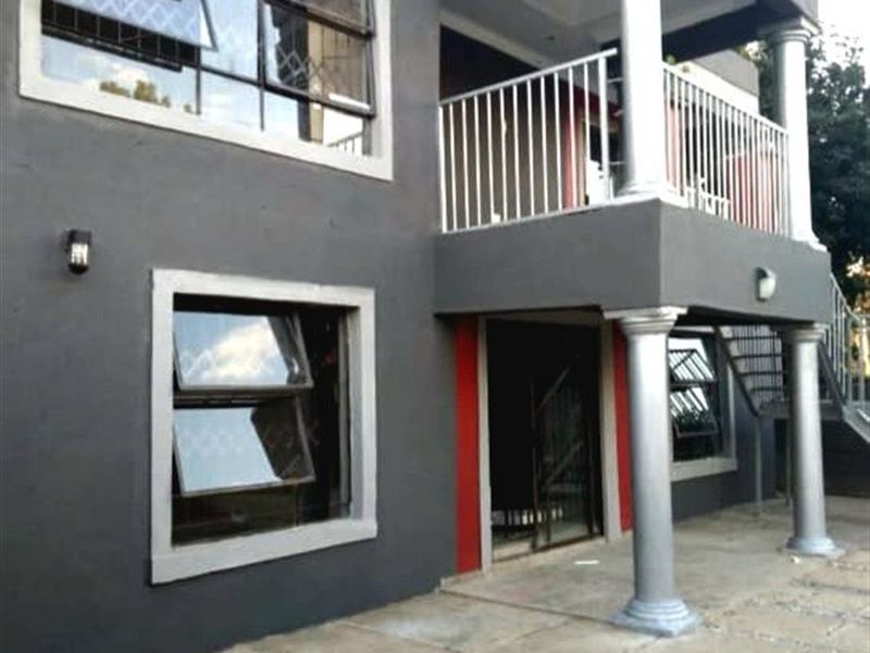 Two-story apartment exterior featuring a paved patio, upper-level balcony with metal balustrade, and modern grey facade with decorative columns.