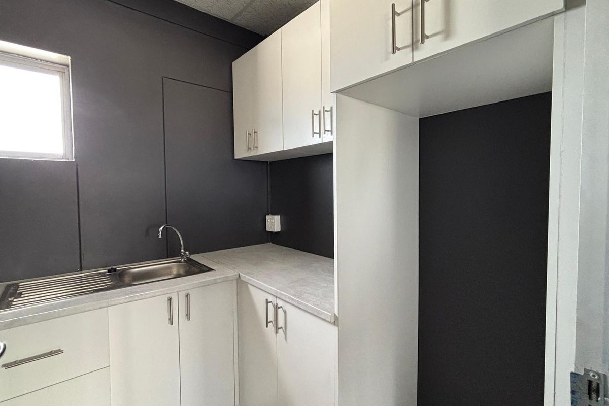 A compact kitchenette featuring modern white cabinetry, a stainless steel sink, and a dedicated space for a refrigerator against a dark accent wall.