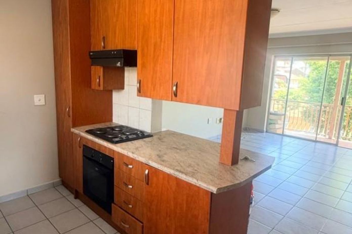 A functional kitchen featuring wooden cabinetry, built-in oven, electric hob, and tiled flooring, opening into a bright living area with balcony access.