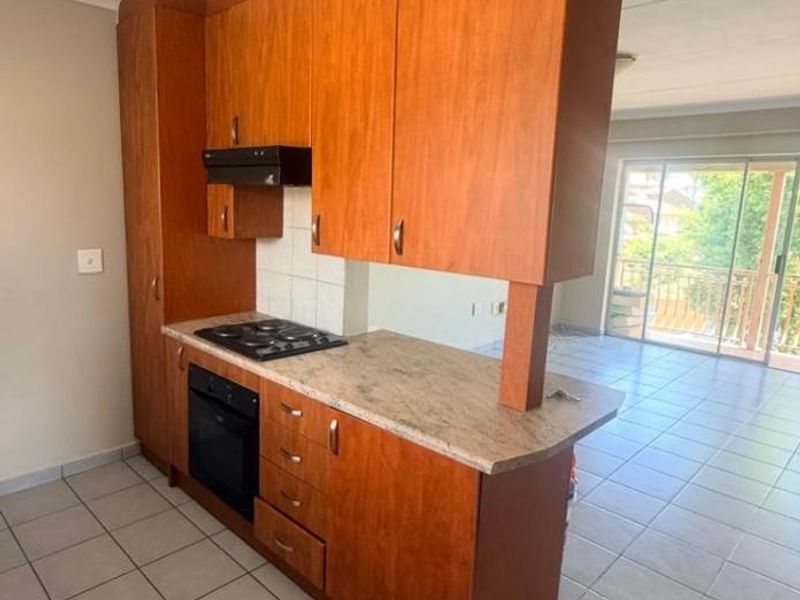 A functional kitchen featuring wooden cabinetry, built-in oven, electric hob, and tiled flooring, opening into a bright living area with balcony access.