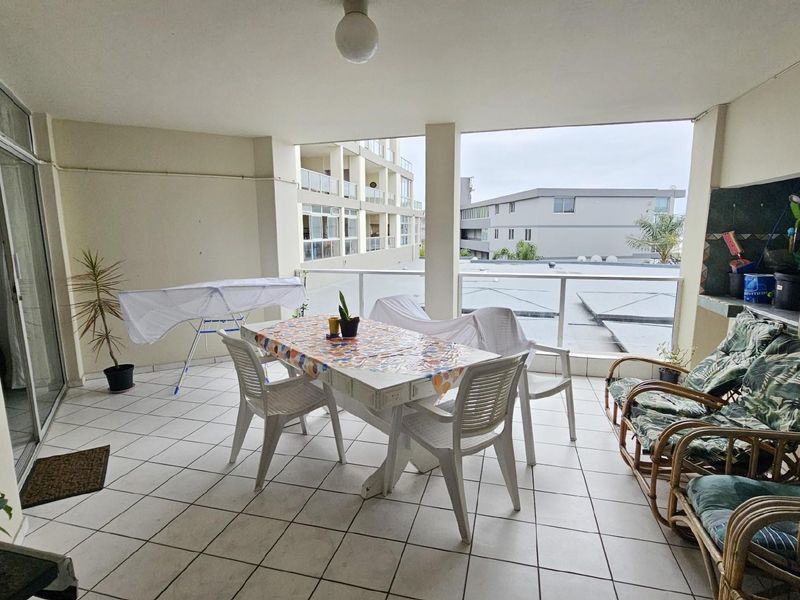 A spacious covered balcony featuring tiled flooring, a built-in braai area, and glass balustrades, offering an outdoor living space with neighbourhood views.