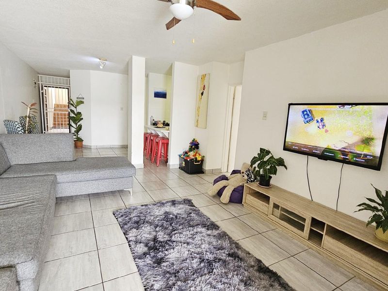 Open plan living area featuring tiled flooring, a ceiling fan, and a wall-mounted television setup, flowing into a kitchen area with a breakfast bar.
