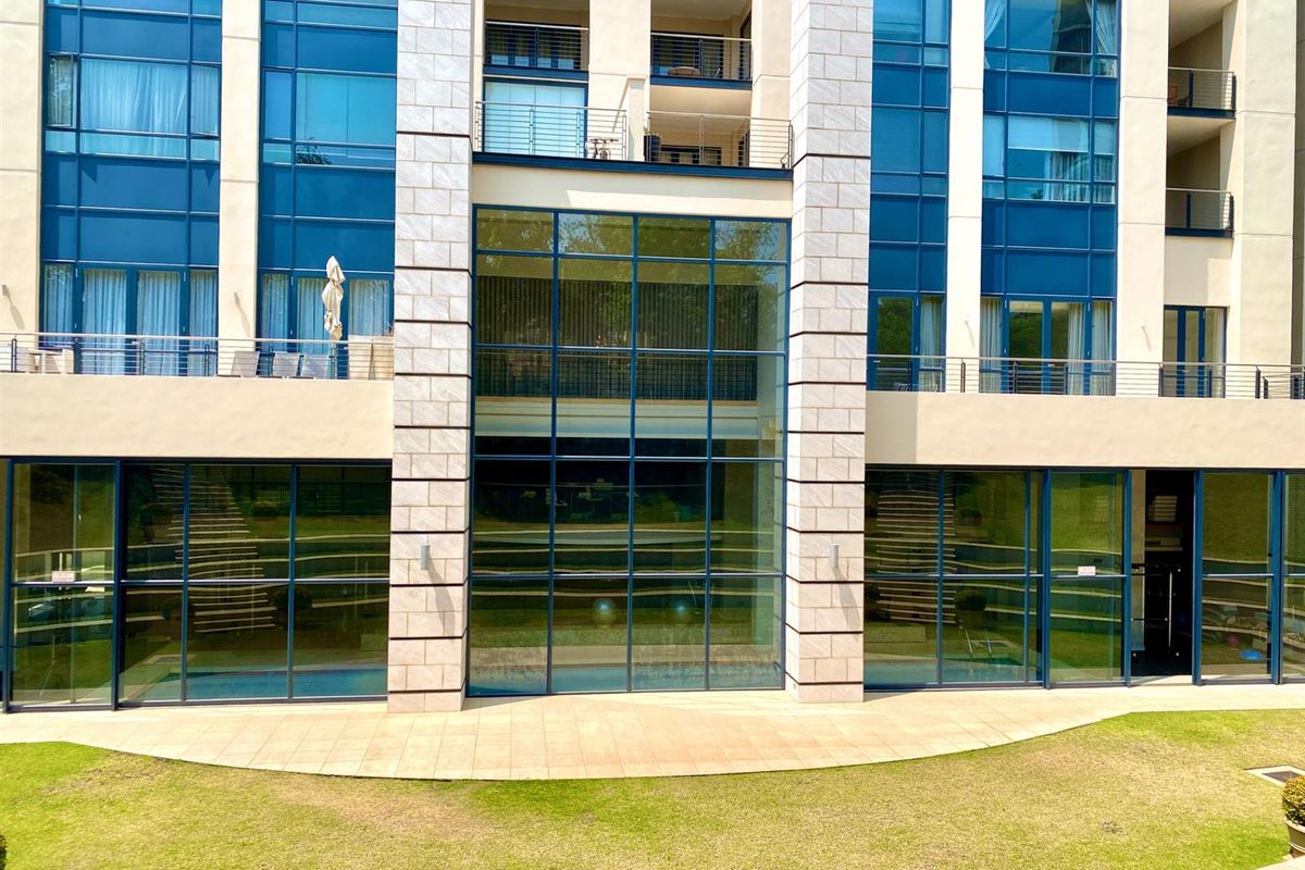 A view of the modern apartment building facade featuring large glass curtain walls, stone-clad pillars, and balconies overlooking a landscaped garden area.