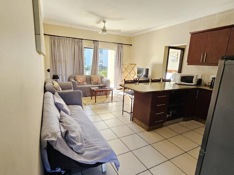 Open-plan living area featuring a kitchen with dark wood cabinetry, breakfast bar, and tiled flooring, flowing into a bright lounge with balcony access.