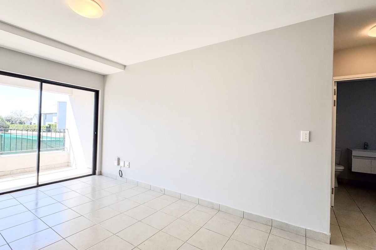 A bright, empty bedroom featuring tiled flooring, a large sliding glass door leading to a balcony, and direct access to an en-suite bathroom.