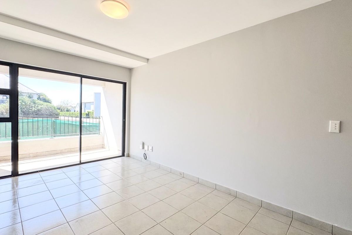 A bright, empty living room featuring neutral ceramic floor tiles and a large sliding glass door providing access to an outdoor balcony area.