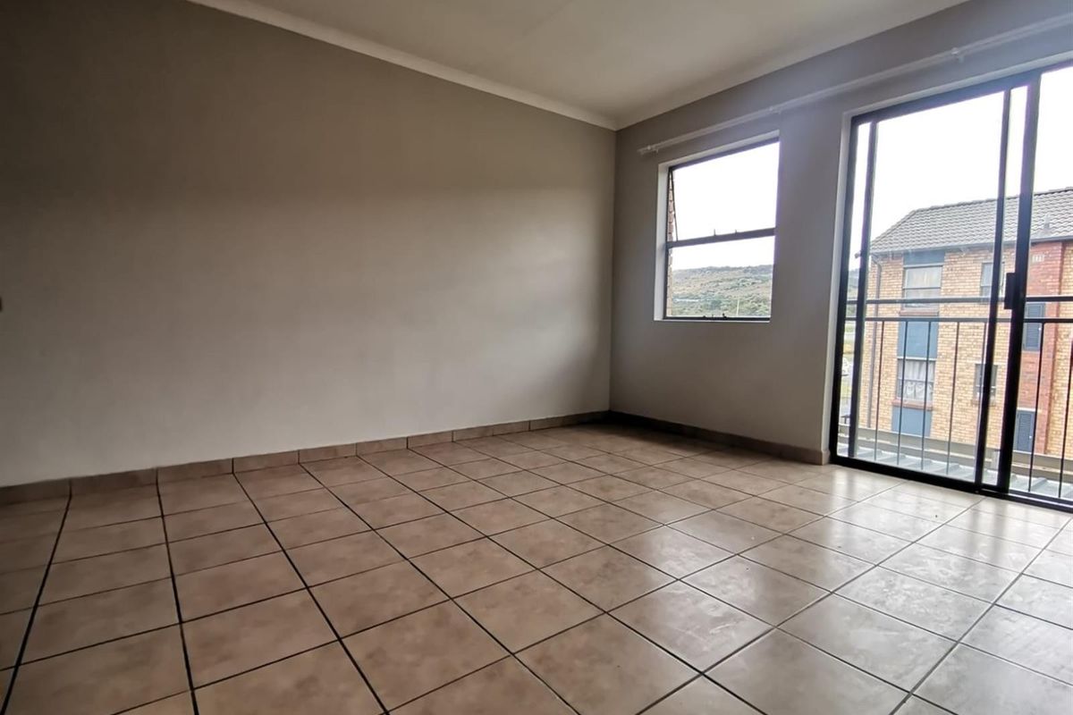 An empty living room featuring neutral tiled flooring, a sliding glass door leading to a balcony, and a window providing natural light.