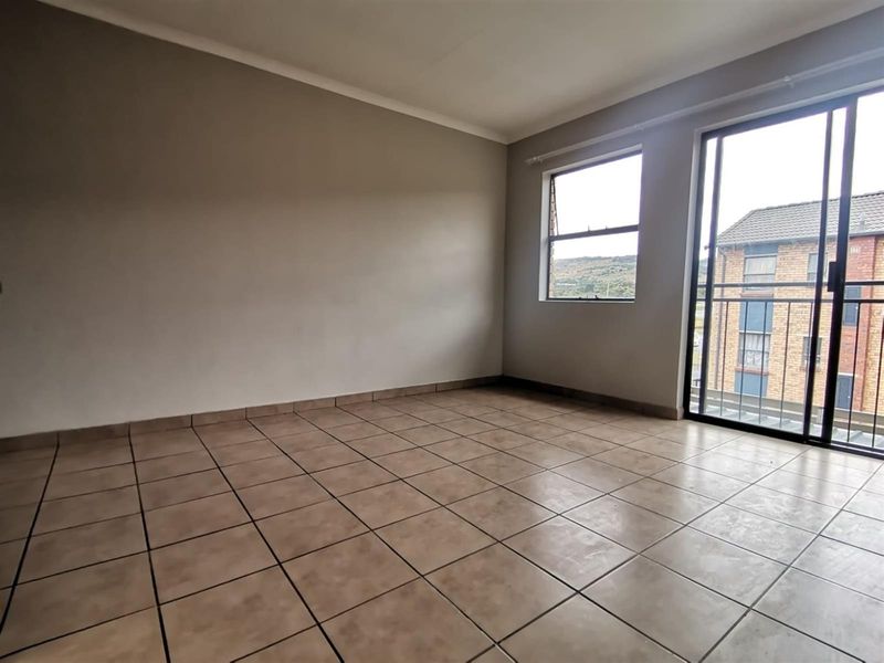 An empty living room featuring neutral tiled flooring, a sliding glass door leading to a balcony, and a window providing natural light.