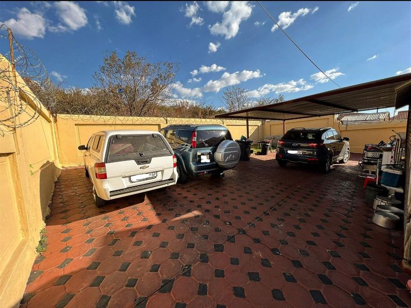 A paved outdoor parking area featuring a covered carport structure, enclosed by high perimeter walls with security razor wire.