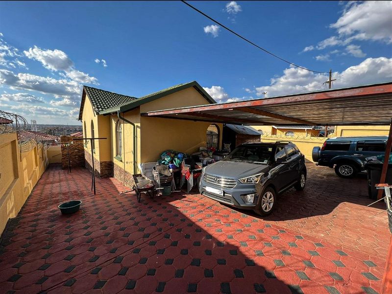 Rear exterior view of the property featuring a paved driveway, a covered carport, and perimeter walling with security fencing.