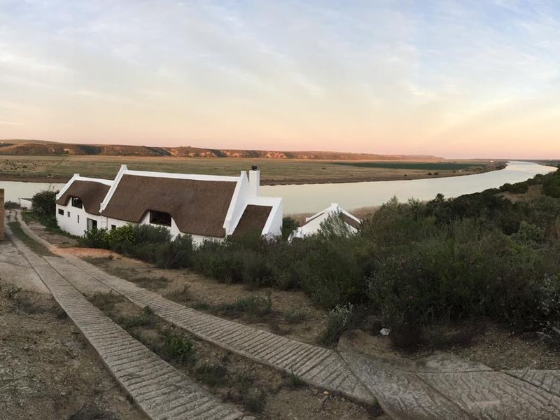 Panoramic view of a traditional thatched-roof farmhouse situated along a riverbank, featuring a concrete driveway and expansive rural landscape.