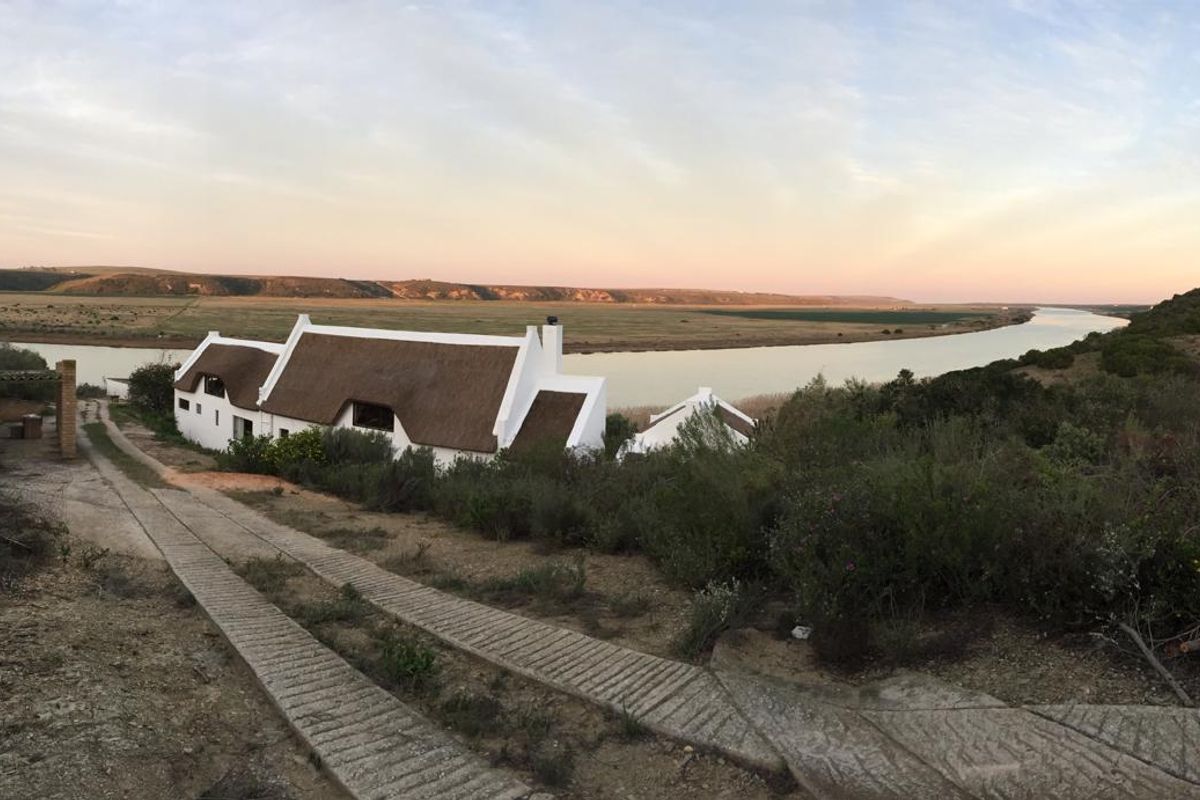 Panoramic view of a traditional thatched-roof farmhouse situated along a riverbank, featuring a concrete driveway and expansive rural landscape.