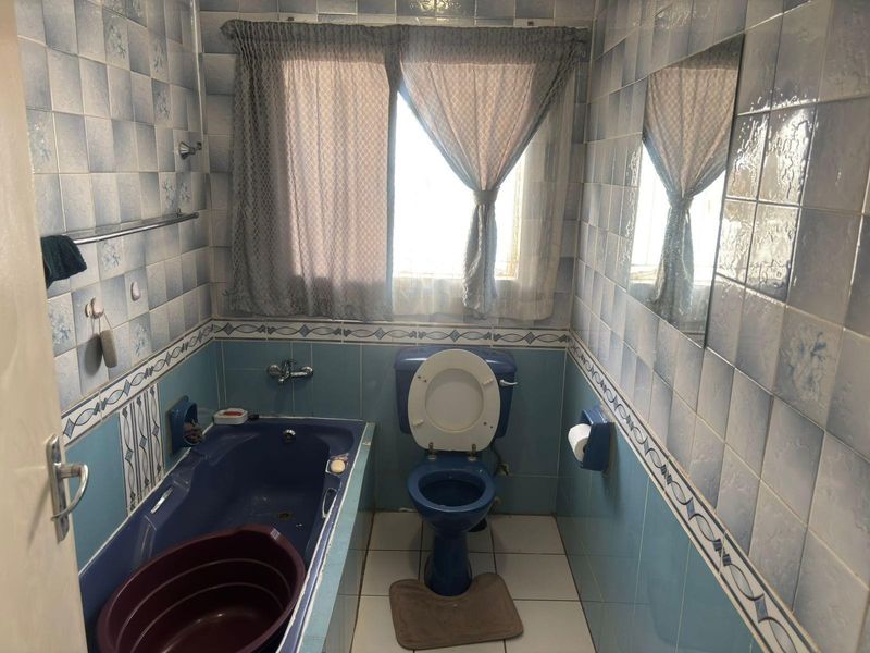 A dated bathroom featuring blue sanitaryware, fully tiled walls, a built-in bathtub, and a window for natural light.