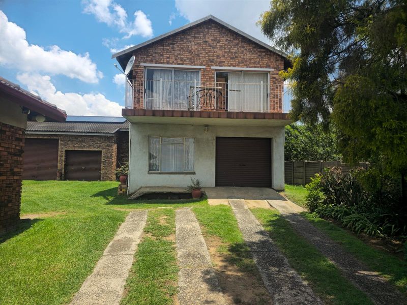 Two-story residential property featuring a brick upper level with a balcony, a ground-level garage, and a concrete driveway set within a garden.