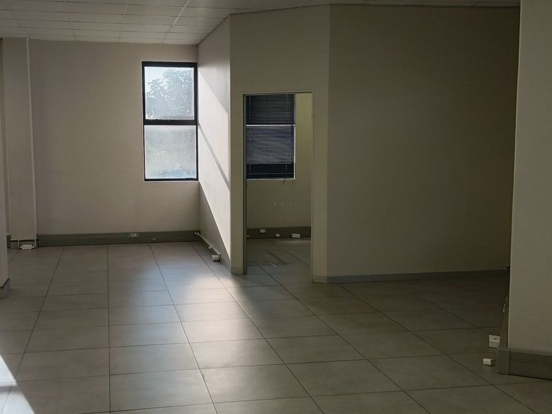 An open-plan office space featuring tiled flooring, a suspended ceiling, and natural light from a side window, leading into a smaller partitioned office area.