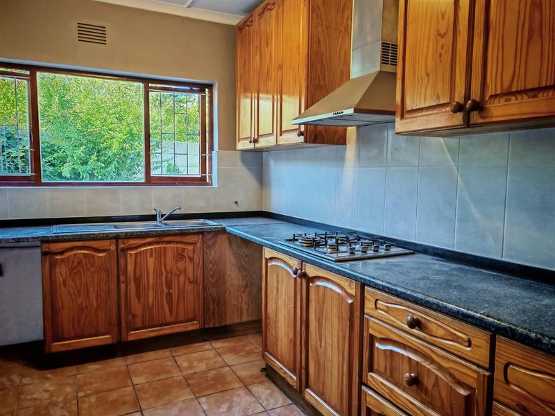 A functional kitchen featuring wooden cabinetry, dark laminate countertops, a gas hob with extractor fan, and tiled flooring with a garden-facing window.