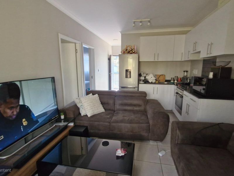 Open-plan living area featuring a compact kitchen with white cabinetry, tiled floors, and integrated appliances, seamlessly connecting to the lounge space.