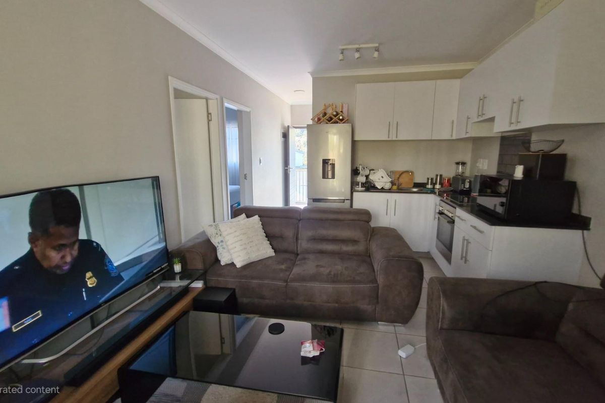 Open-plan living area featuring a compact kitchen with white cabinetry, tiled floors, and integrated appliances, seamlessly connecting to the lounge space.