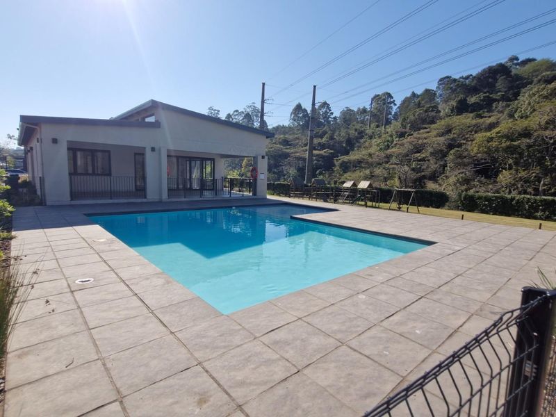 A well-maintained outdoor swimming pool area featuring a paved deck, metal fencing, and an adjacent covered patio space within a landscaped setting.