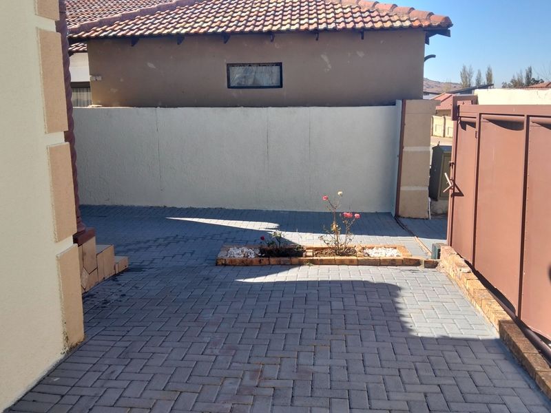 A paved front driveway area featuring a boundary wall, metal gate, and a small central garden bed.