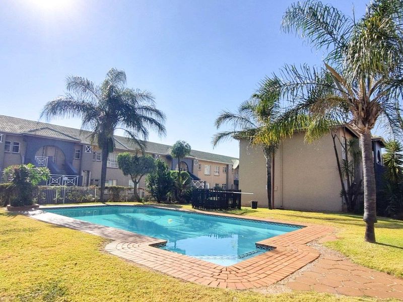 Communal outdoor area featuring a swimming pool with brick paving, surrounded by landscaped lawns and established palm trees within the apartment complex.