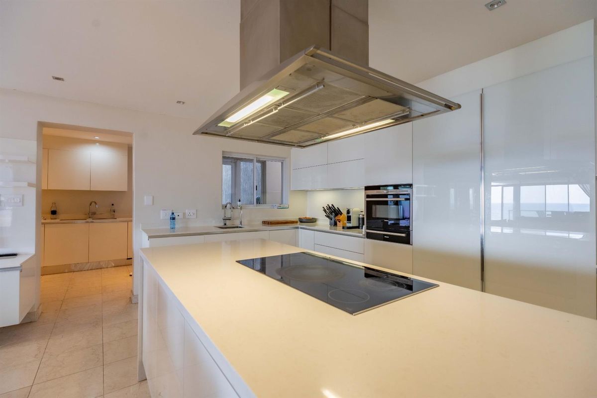 Modern kitchen featuring a central island with a built-in electric cooktop, overhead extractor fan, and sleek white cabinetry, with an adjacent scullery area.
