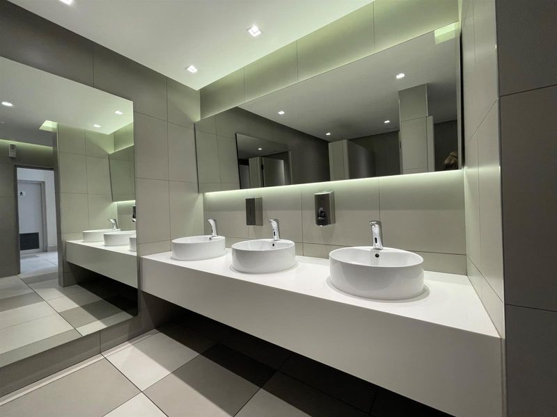 Modern office bathroom featuring a sleek triple vessel sink vanity, large wall mirror, recessed lighting, and contemporary neutral tiling throughout.