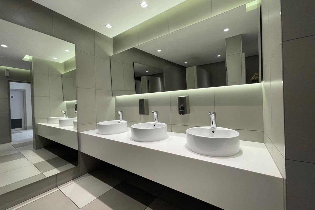 Modern office bathroom featuring a sleek triple vessel sink vanity, large wall mirror, recessed lighting, and contemporary neutral tiling throughout.