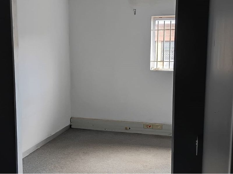A small, empty office space featuring carpeted flooring, a window with security bars, and wall-mounted electrical outlets.