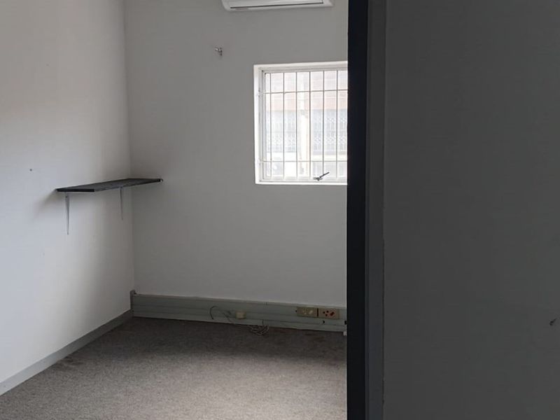A small, empty office space featuring carpet flooring, a wall-mounted shelf, security bars on the window, and an air conditioning unit.