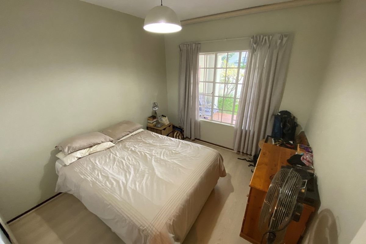 A functional bedroom featuring light-toned laminate flooring, a large window with curtain tracks, and a central pendant light fixture.