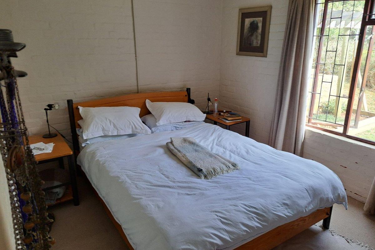 A cozy bedroom featuring painted brick walls, carpeted flooring, and a large window with security bars overlooking the garden.
