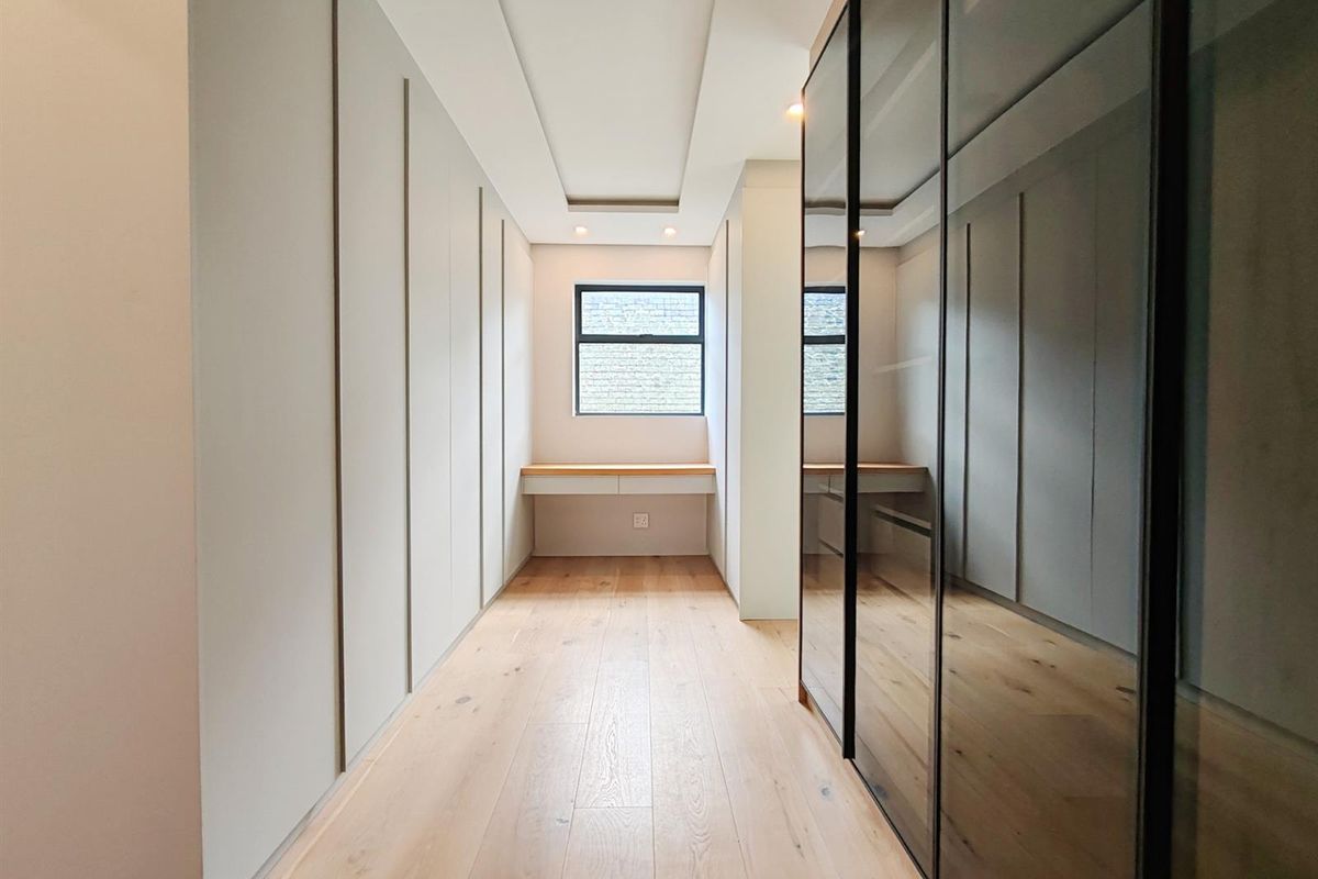A modern walk-in closet featuring extensive floor-to-ceiling built-in cabinetry, integrated glass-fronted wardrobes, light timber flooring, and a dedicated built-in vanity desk.