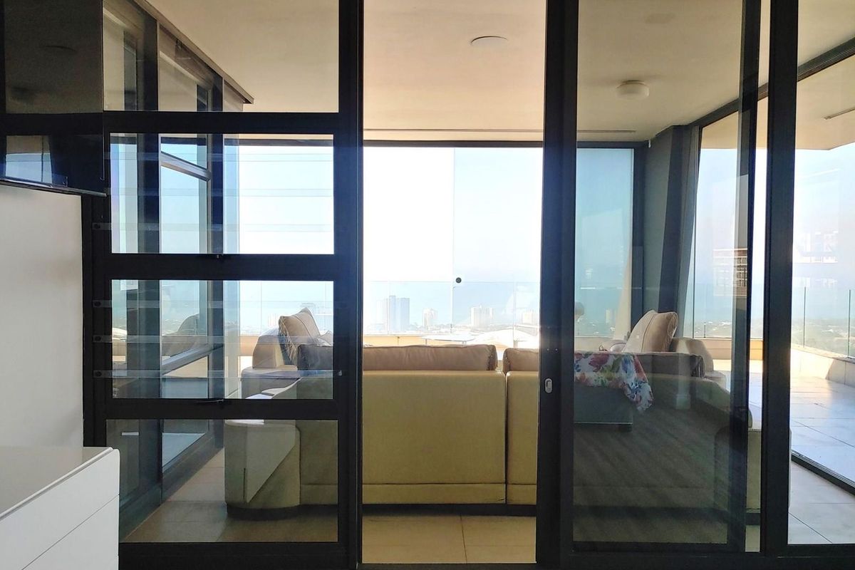 A bright living area featuring floor-to-ceiling glass sliding doors that open onto a spacious balcony with expansive city and coastal views.